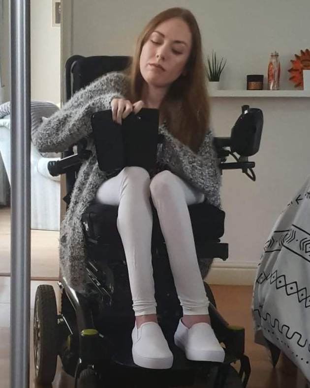 A selfie of me, looking in the mirror, seated in my powered wheelchair. I have long, mid-brown hair and I'm wearing a grey cardigan and white leggings. In this photo, I am aged 33